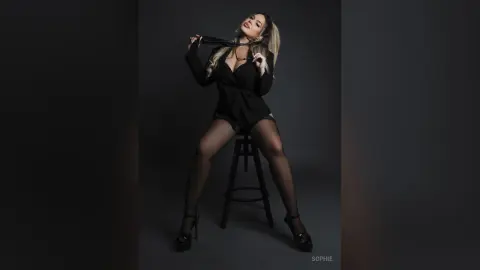 Elegant woman in a black blazer and sheer tights posing on a stool against a dark studio background.
