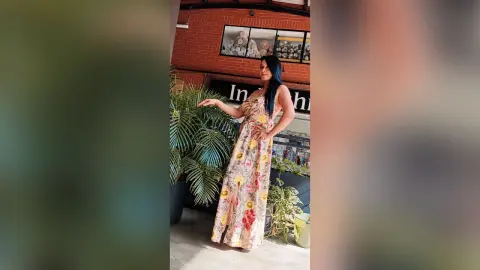 MeganneAllen poses outdoors in a long, floral print sundress with a high slit, showing her legs and dark hair.