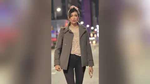 HaleyLyon wears a grey coat over a light lace top and black pants while standing on a city street at night.