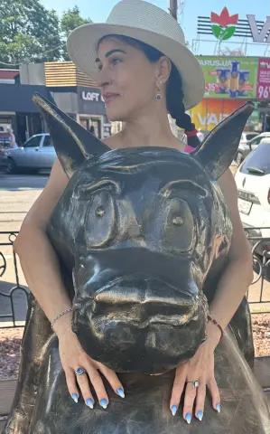 Smiling woman in a white sun hat leaning against a large bronze bull statue on a sunny city street.