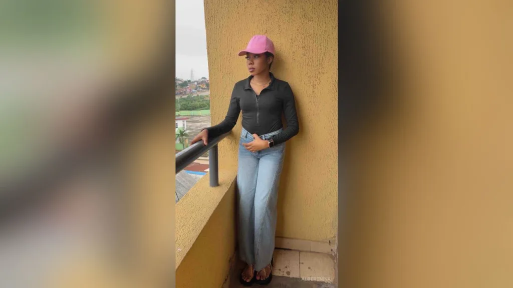 AlbertNina — AlbertNina stands on a balcony wearing a black long-sleeve shirt, blue jeans, and a pink cap.