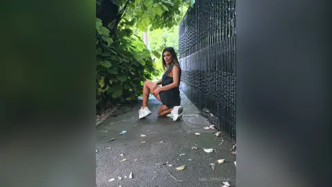 Young woman in a black mini dress and white sneakers kneeling on a leafy urban path by a black metal fence.
