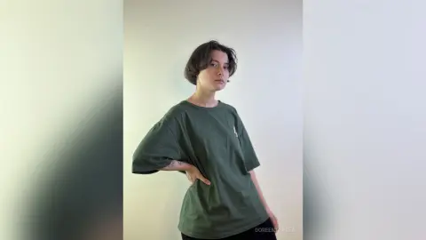 Young man in an oversized dark green t-shirt posing against a minimalist white wall with a relaxed mood.