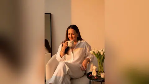 Relaxed woman in white linen loungewear sipping coffee in a warm, cozy indoor setting.