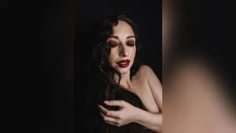 Elegant woman with wavy dark hair and bold red lipstick posing in a dark, moody setting.