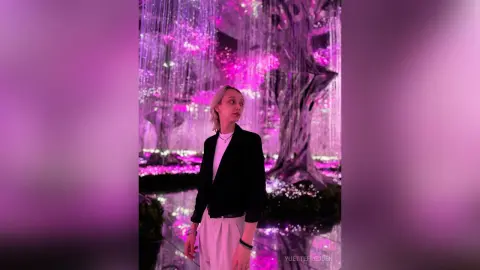 Blonde woman in a black blazer and white top standing in a dreamy, glowing pink neon light installation.