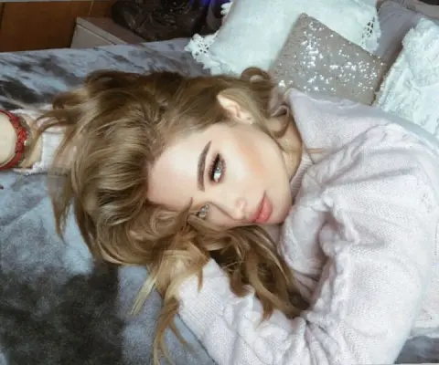Blonde woman in a cozy white cable-knit sweater relaxing on a bed with soft pillows.