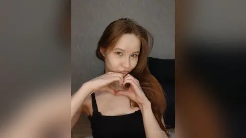 Young woman with long red hair in a black tank top making a heart gesture against a neutral gray background.