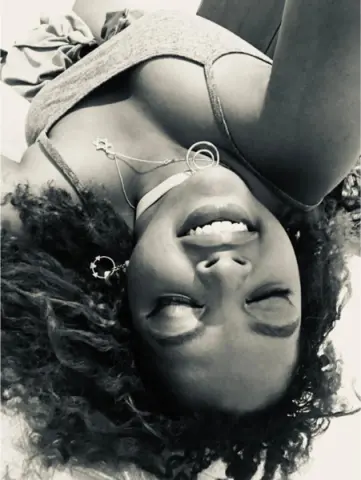 Smiling Black woman with curly hair wearing a grey tank top and silver necklace in a bright, joyful setting.