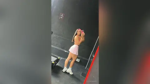 Fit woman in pink crop top and skirt posing in a mirror at a modern dark gym.