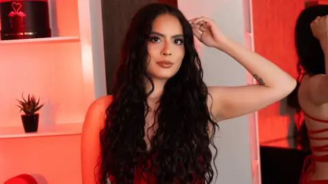 Woman with long wavy dark hair in a red outfit posing in a warm, red-lit room.