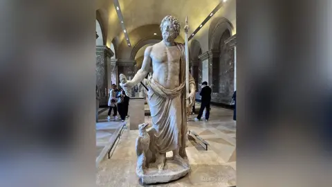 Ancient Greek marble statue of a bearded man holding a spear in a grand museum hall.