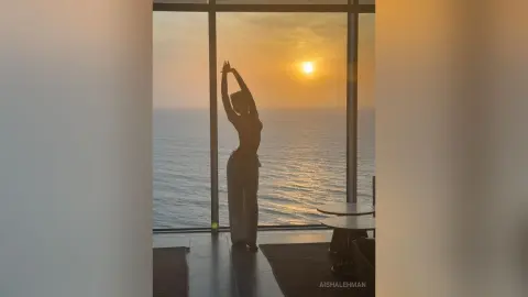 Silhouette of a woman stretching by a high-rise window overlooking a golden ocean sunset.