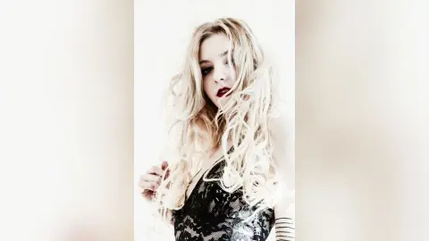 Blonde woman in a black lace outfit posing with a dreamy, ethereal mood against a white background.