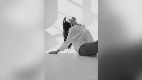 Dreamy black and white portrait of a woman in a white button-down shirt leaning back against a bright wall.