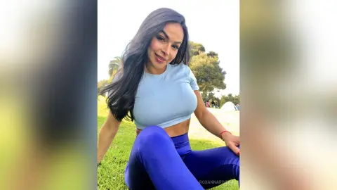 Smiling woman in blue crop top and leggings relaxing on green grass in a sunny park.