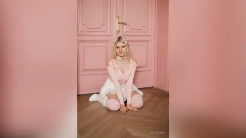Blonde woman in a pink fairy outfit sitting on a wooden floor against a soft pink door background.