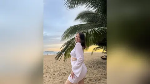 EmorySwift smiles while posing on a sandy beach, wearing a white long-sleeve crop top and matching white shorts under a palm tree.