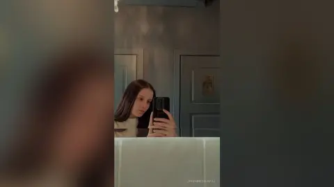 Young woman in a white top taking a moody mirror selfie in a dimly lit room.