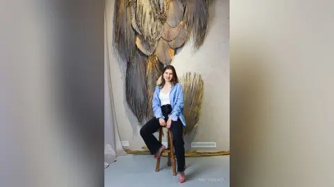 Young woman in a blue shirt and black pants sitting on a stool against a large feather wall art backdrop.