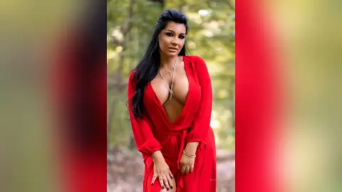 MissyJolie poses in a red plunging neckline dress with her long black hair down in a wooded outdoor setting.