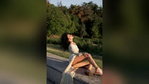 Curvy woman in a white flowing dress sitting on a stone ledge in a lush green forest during golden hour.