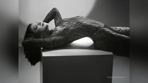 Elegant woman in a black lace bodysuit reclining on a pedestal in a moody black and white studio setting.