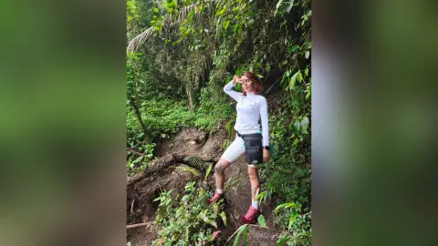 AmelieLautrec stands in a lush green jungle wearing a white long-sleeve shirt, white shorts, and a black waist bag, shielding her eyes with one hand.