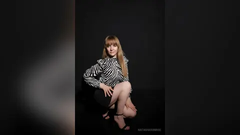 NatashaSunrise — NatashaSunrise poses in a black and white striped blouse and black skirt against a dark background.