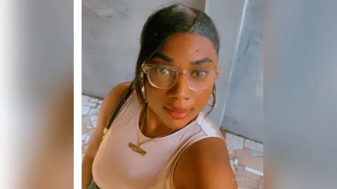 MandyJohn — MandyJohn wears glasses and a beige tank top while posing for a selfie outdoors.