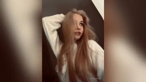 AletaKunde poses with her long blonde hair, wearing a white long-sleeved shirt against a dark background.