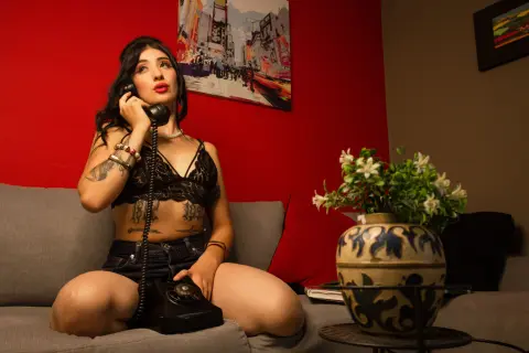 Woman in black lace lingerie and denim shorts talking on a vintage telephone in a moody red room.