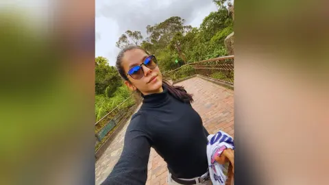 JessieLiu wears a black turtleneck and blue-tinted sunglasses while standing on a brick walkway outdoors near green trees.