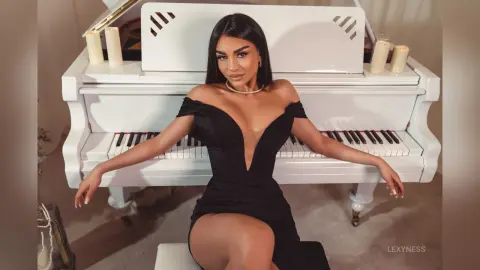 Elegant woman in a black plunging evening gown sitting by a white grand piano in a sophisticated indoor setting.
