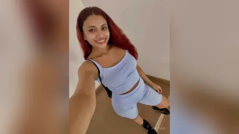 Smiling woman in a light blue ribbed tank top and matching shorts posing indoors for a casual selfie.