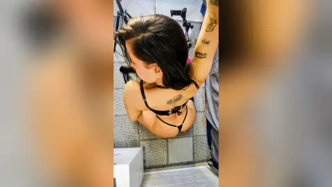 ElizaLuxe poses with her back to the camera, showing her bare back, tattoos, and black thong while leaning forward on a grey floor.