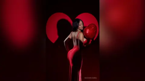 NohomiCambel poses from behind in a red dress, looking over her shoulder against a red heart-shaped background.