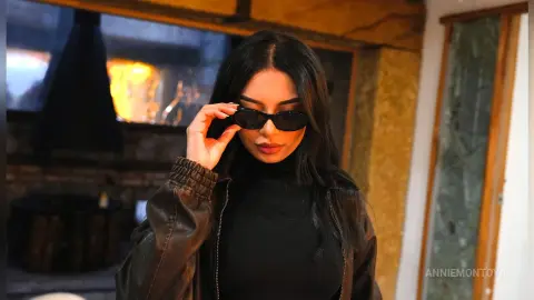 Stylish woman in a black turtleneck and leather jacket wearing sunglasses in a cozy indoor setting.