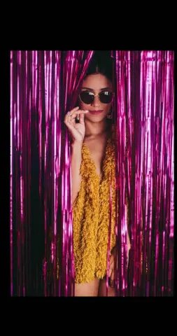 CelesteGrayy poses in a fuzzy yellow mini dress and sunglasses behind pink tinsel curtains.