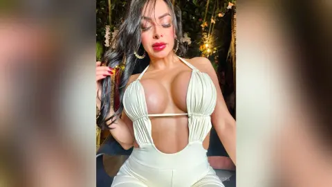Glamorous woman in a white plunging halter jumpsuit posing in a lush, dimly lit garden setting.