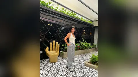 Stylish woman in a white tank top and grey trousers standing by a gold hand sculpture in a modern patio.
