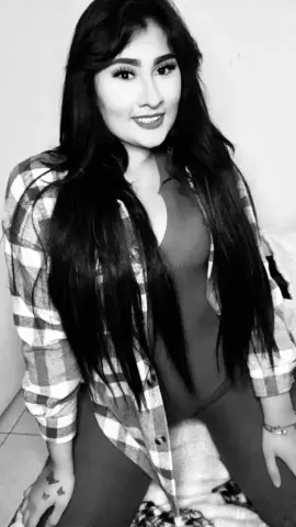 Smiling young woman with long dark hair wearing a plaid shirt and dark top in a cozy indoor setting.