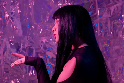 Young woman with long black hair in a black cutout top posing against a shimmering pink glitter background.
