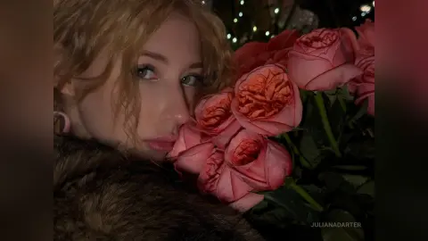 Blonde woman in a brown faux fur coat posing with a large bouquet of pink roses in soft evening lighting.