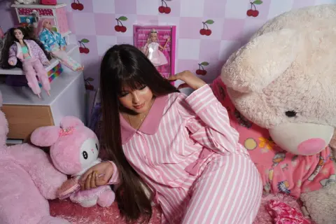 Young woman in pink striped pajamas relaxing in a dreamy pink bedroom surrounded by plush teddy bears and dolls.