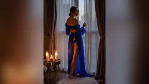 Elegant woman in a blue sheer robe standing by a window next to glowing candles in a moody, romantic room.
