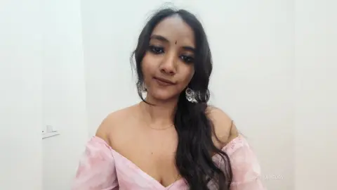 Young woman with long dark hair wearing a pink off-the-shoulder top and silver earrings in a bright room.