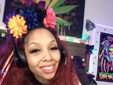 Tasha Vegas — Tasha Vegas smiles wearing a colorful flower crown and black headphones against a dark background with weed leaf art.