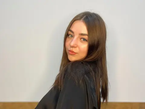 Lois Below — Lois Below looks over her shoulder with long brown hair and a black top against a white wall.