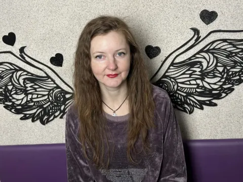 Megan Pirs — Megan Pirs with long wavy hair wears a purple velvet sweatshirt while sitting in front of a winged wall decoration.